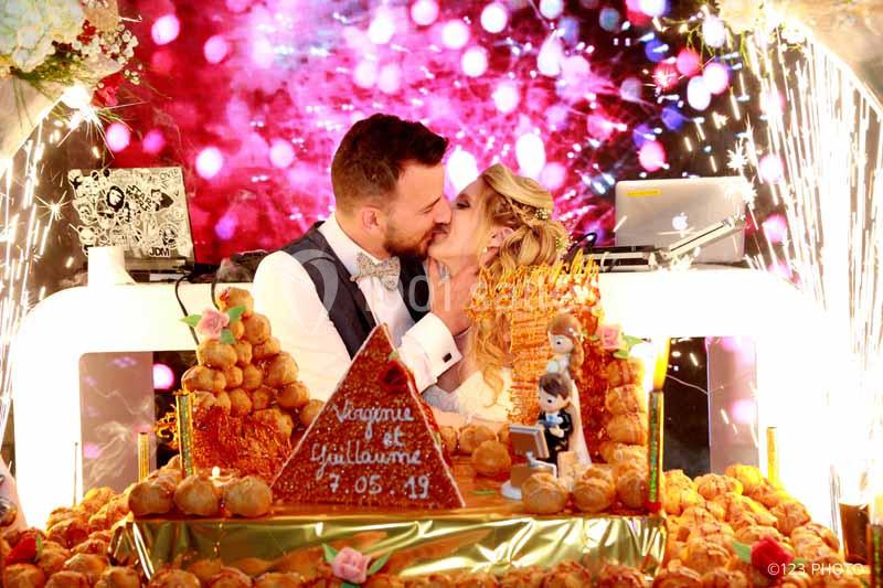 Un couple s'embrasse devant une pièce montée de choux décorée, avec un fond lumineux festif.