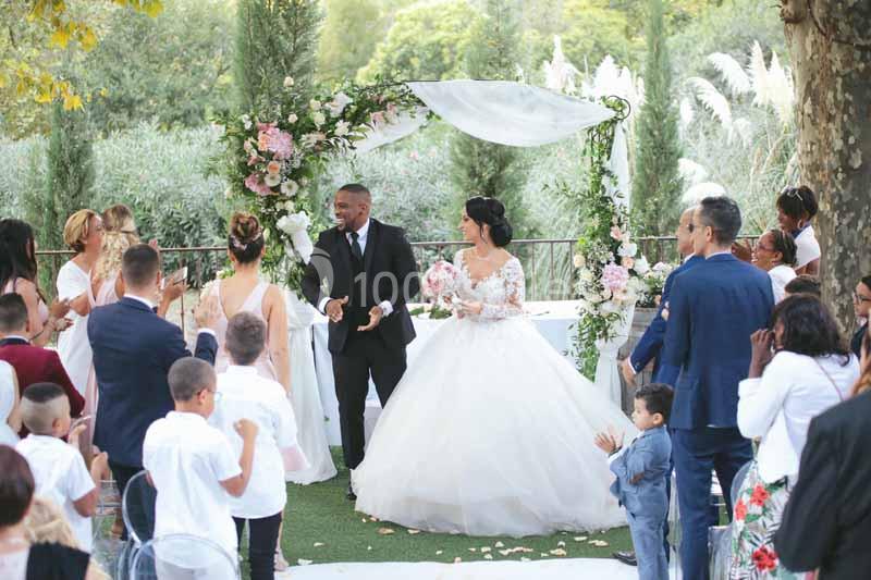Un couple en tenue de mariage marche sous une arche fleurie, entouré d'invités dans un jardin.