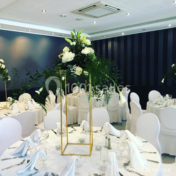 Salle de réception décorée avec des tables rondes, nappes blanches et centre de table floral sur structure dorée.