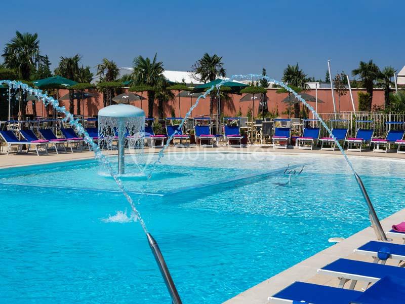 Piscine extérieure avec jets d'eau, transats bleus et parasols entourés de palmiers sous un ciel dégagé.