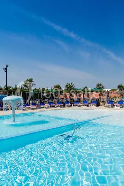 Piscine extérieure avec fontaine, entourée de transats et de parasols sous un ciel bleu dégagé.
