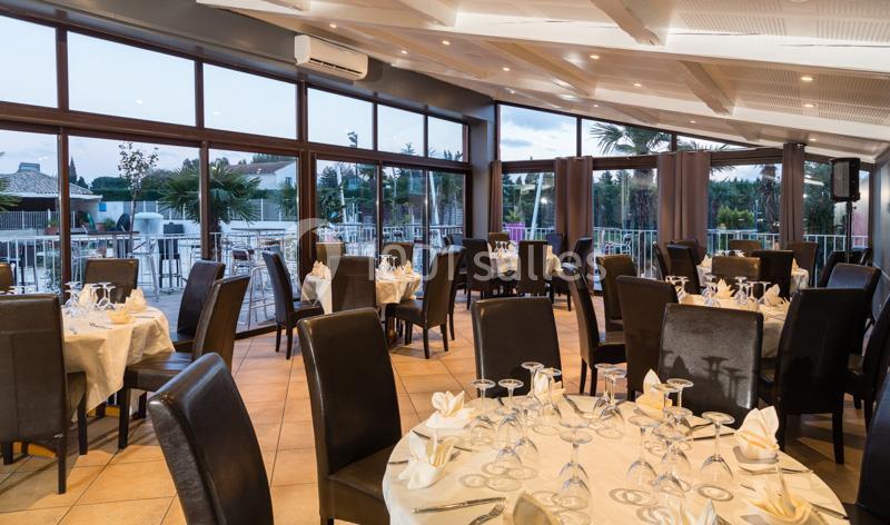 Salle de restaurant lumineuse avec tables dressées, nappes blanches, chaises en cuir sombre et grandes baies vitrées.