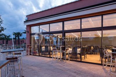 Salle de restaurant lumineuse avec grandes baies vitrées, tables dressées et vue sur une terrasse extérieure.