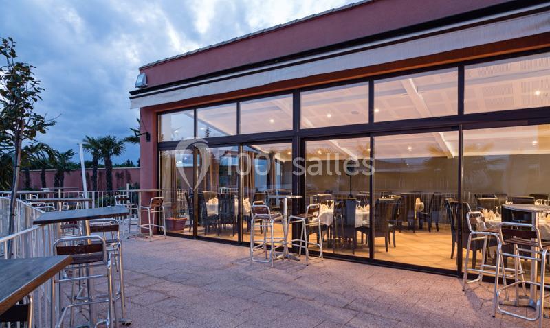 Terrasse extérieure avec tables et chaises devant une grande baie vitrée d'un bâtiment moderne au crépuscule.