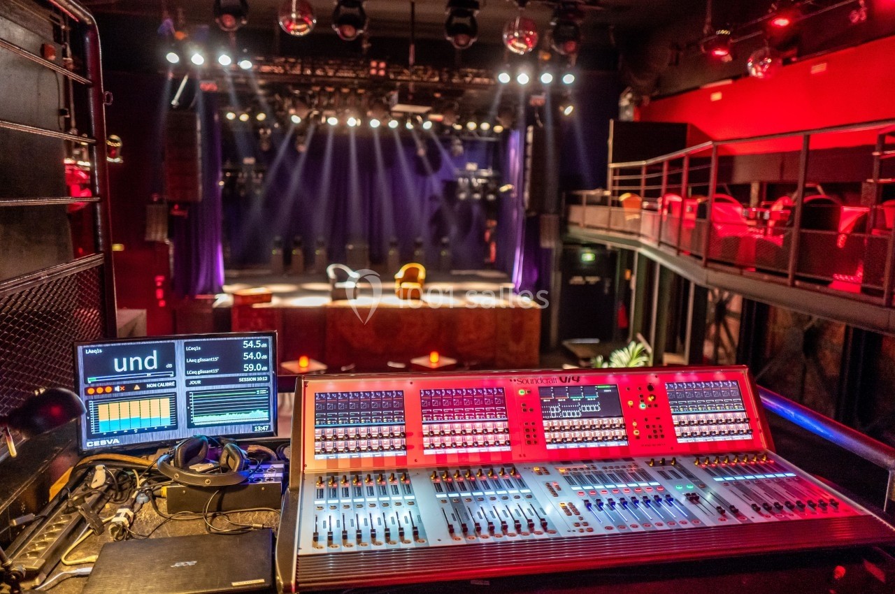 Table de mixage audio avec écran de contrôle en premier plan, face à une scène de théâtre éclairée et des sièges vides.