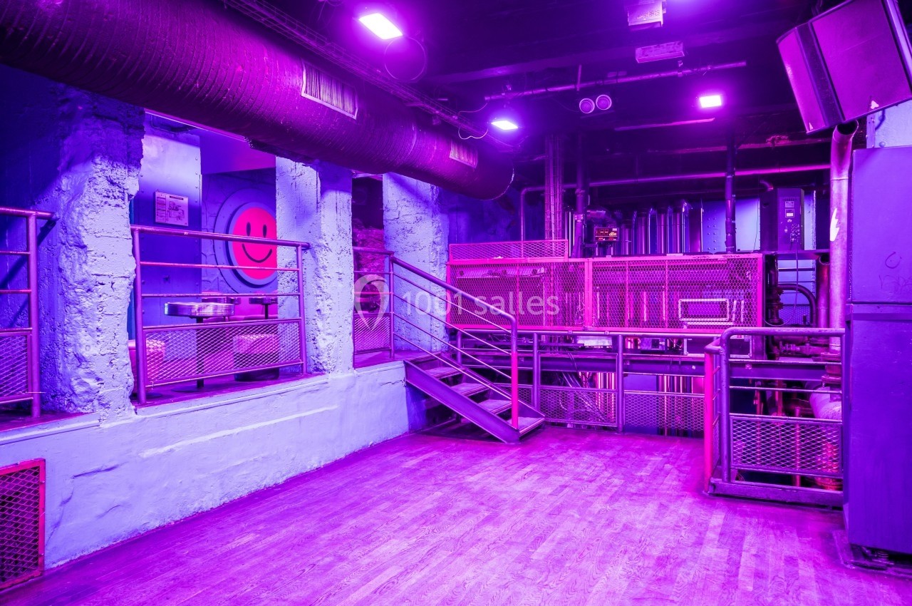 Salle industrielle vide éclairée par une lumière violette, avec des escaliers métalliques et des murs peints en bleu.