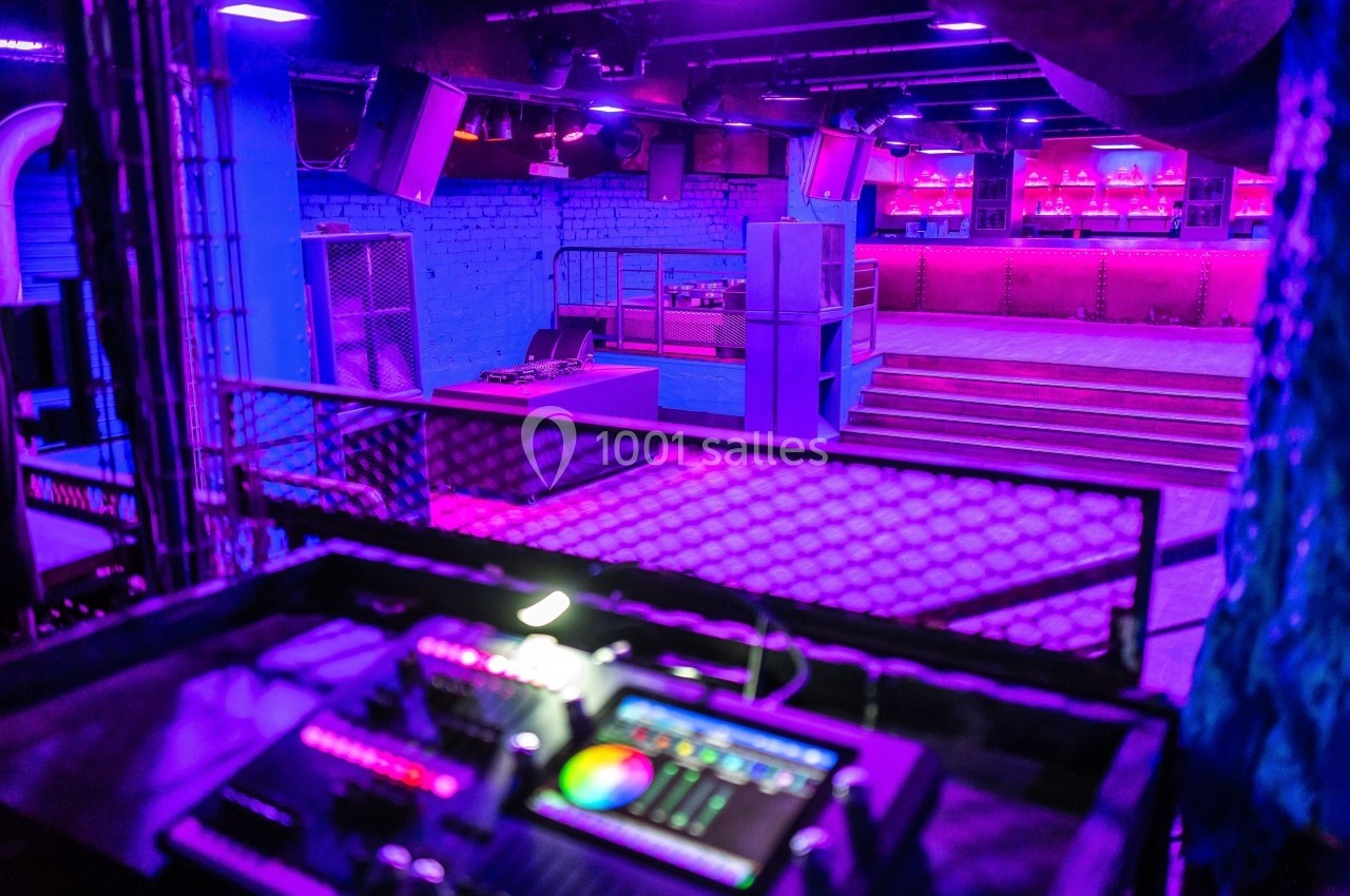 Salle de concert vide avec éclairage violet, une console de mixage au premier plan et un bar en arrière-plan.