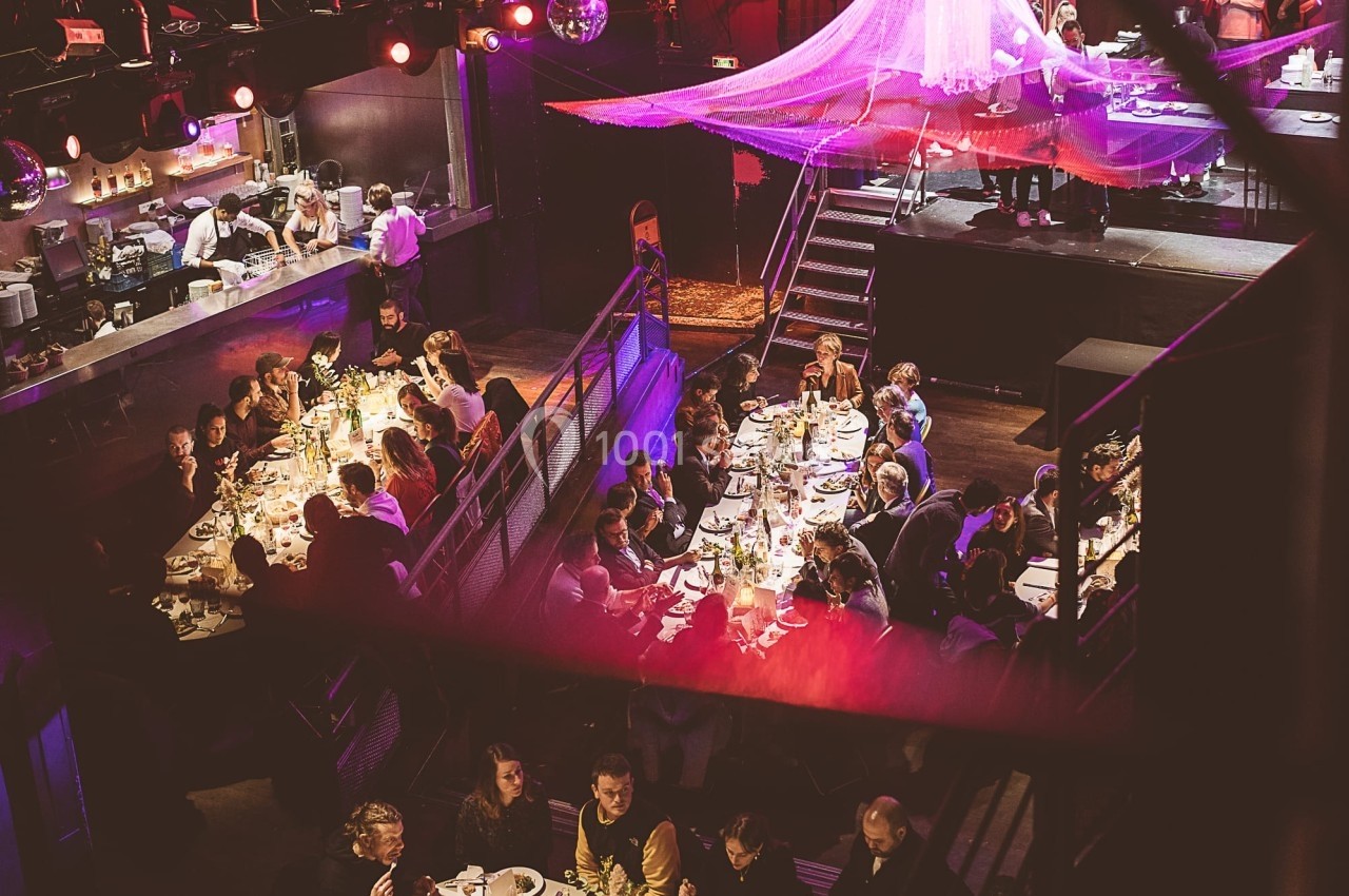 Salle de réception animée avec des convives dînant à des tables éclairées sous une lumière tamisée et colorée.