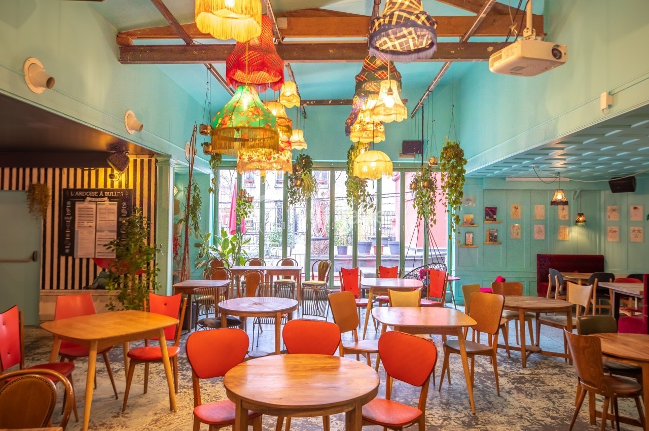 Salle de restaurant colorée avec tables en bois, chaises variées et luminaires suspendus dans un décor lumineux et végétal.