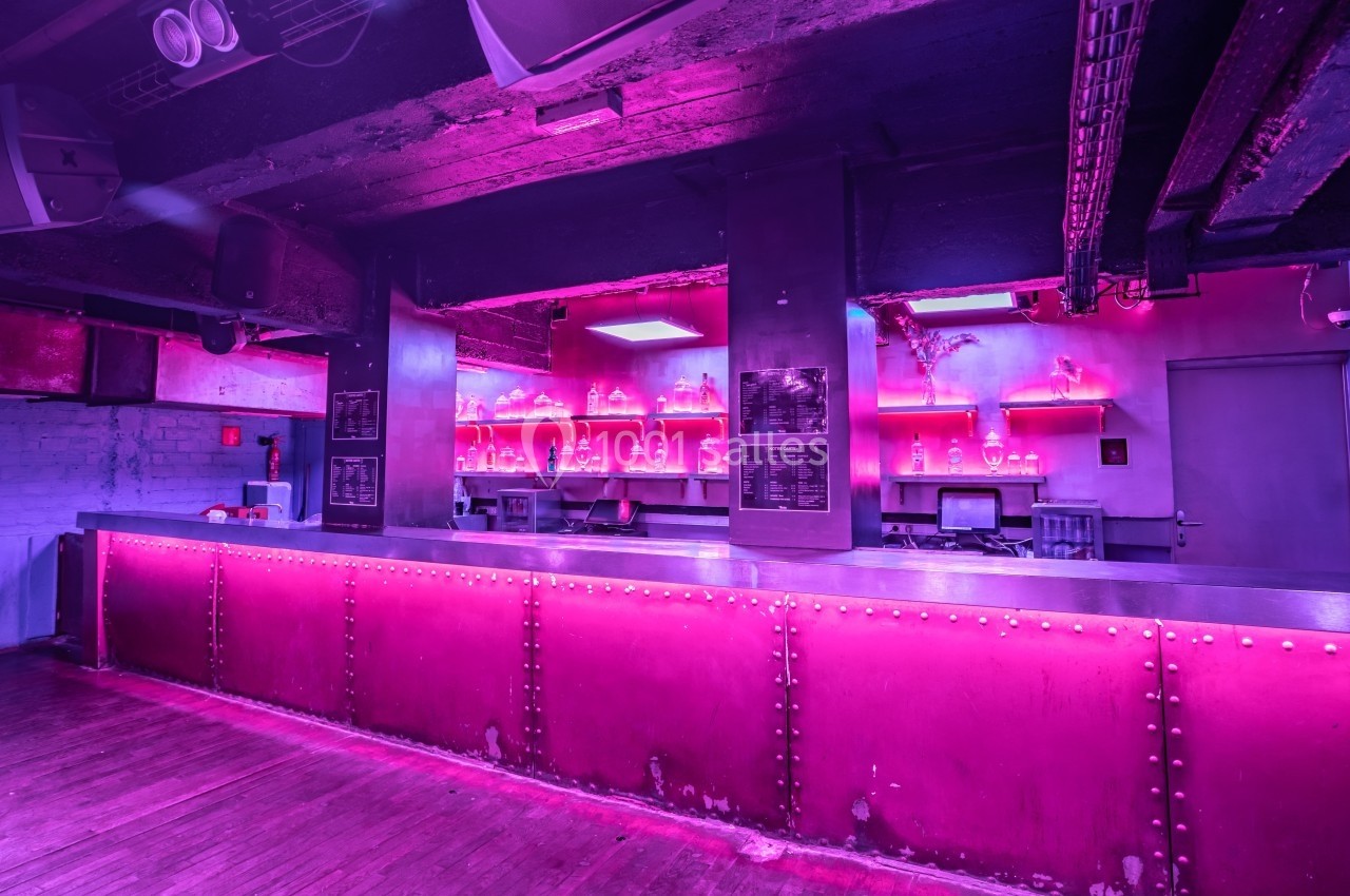 Bar avec comptoir métallique et éclairage rose, étagères avec bouteilles et murs en béton brut dans une ambiance…