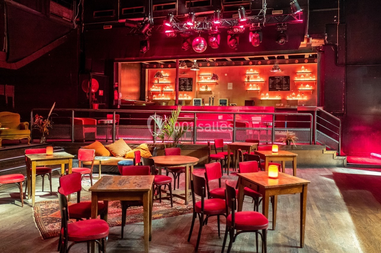 Salle de bar avec tables et chaises en bois, éclairage tamisé rouge et comptoir illuminé en arrière-plan.