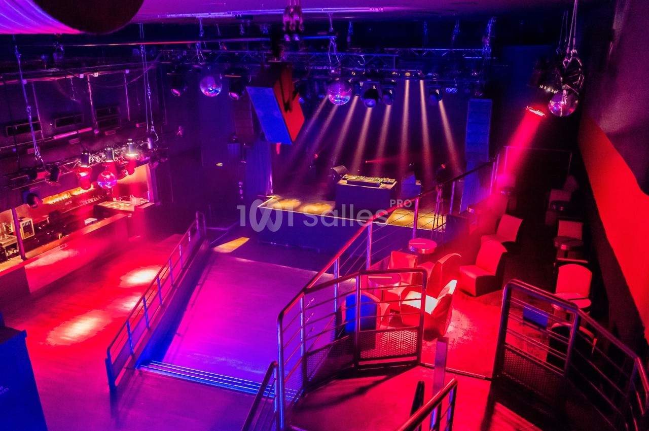 Salle de discothèque vide avec piste de danse, éclairages colorés, tables et scène équipée d'une console DJ.