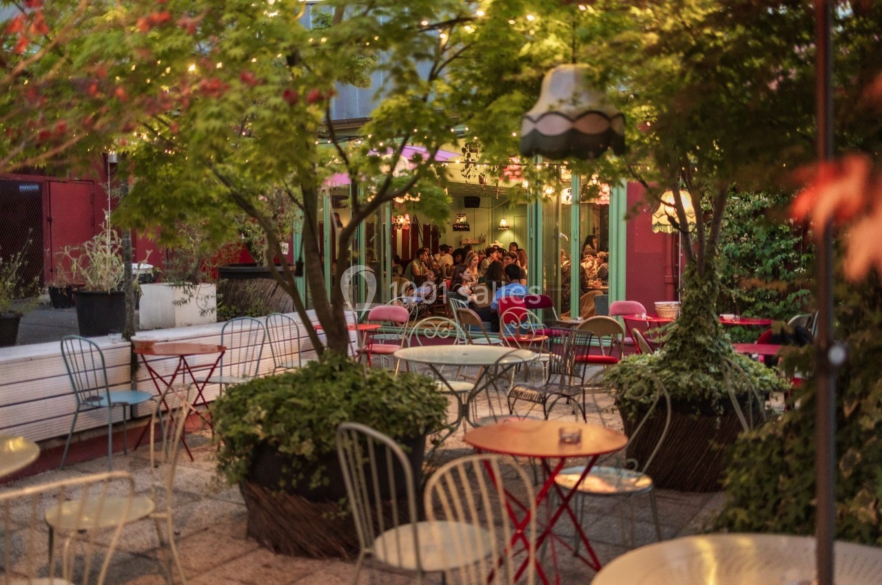 Terrasse extérieure avec tables et chaises entourées de plantes, éclairée par des guirlandes lumineuses en soirée.