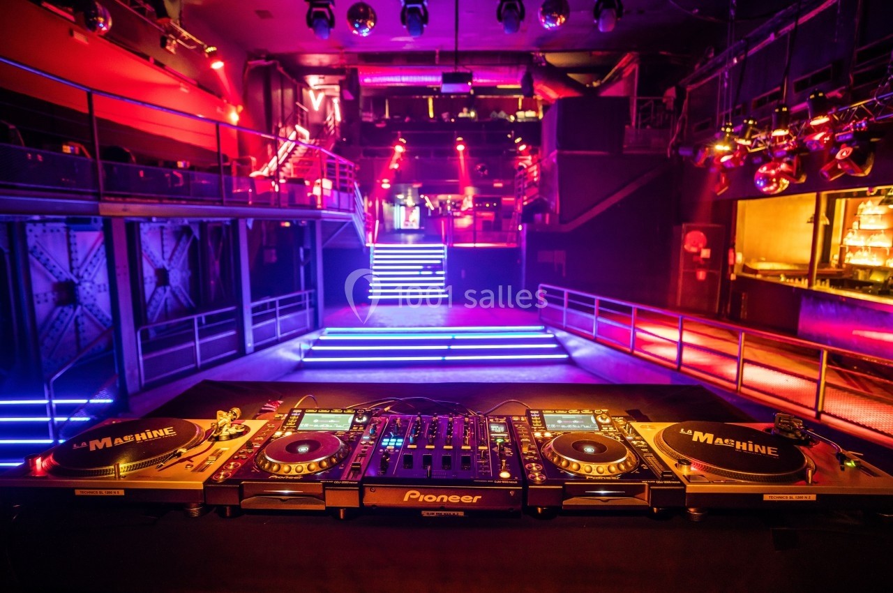 Vue d'une cabine de DJ avec platines et table de mixage dans une salle de concert illuminée par des lumières colorées.