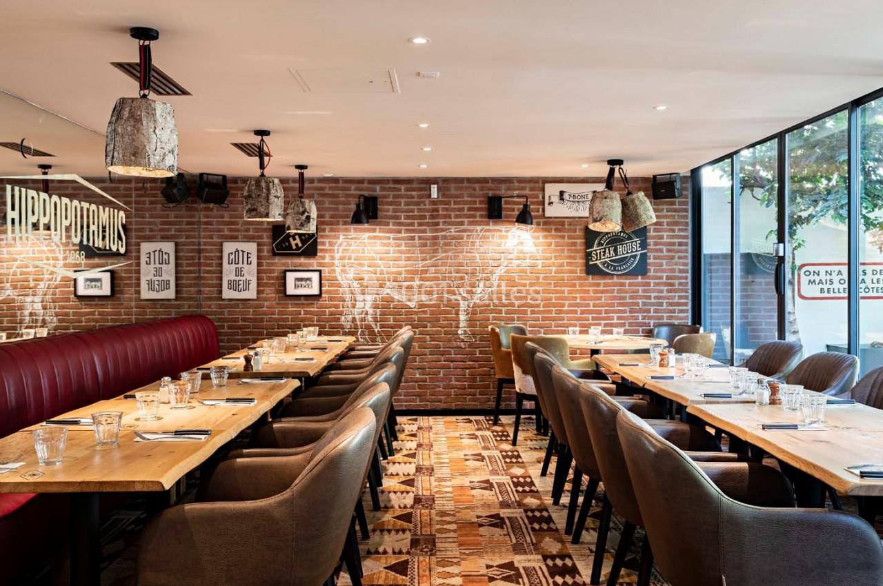 Salle de restaurant avec tables en bois, chaises rembourrées, murs en briques et grande baie vitrée.