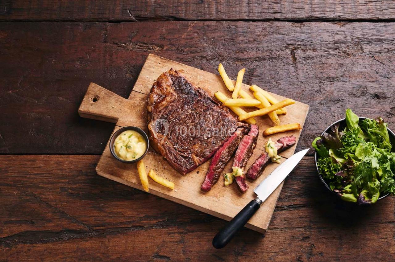 Steak grillé avec frites, sauce et salade, présenté sur une planche en bois avec un couteau sur une table rustique.