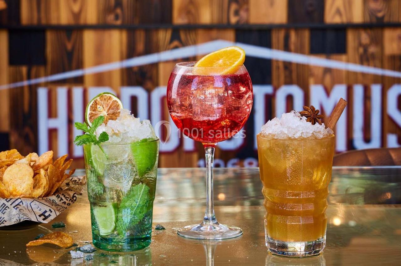 Trois cocktails colorés servis sur une table, accompagnés de chips, avec un fond en bois et une enseigne floue.