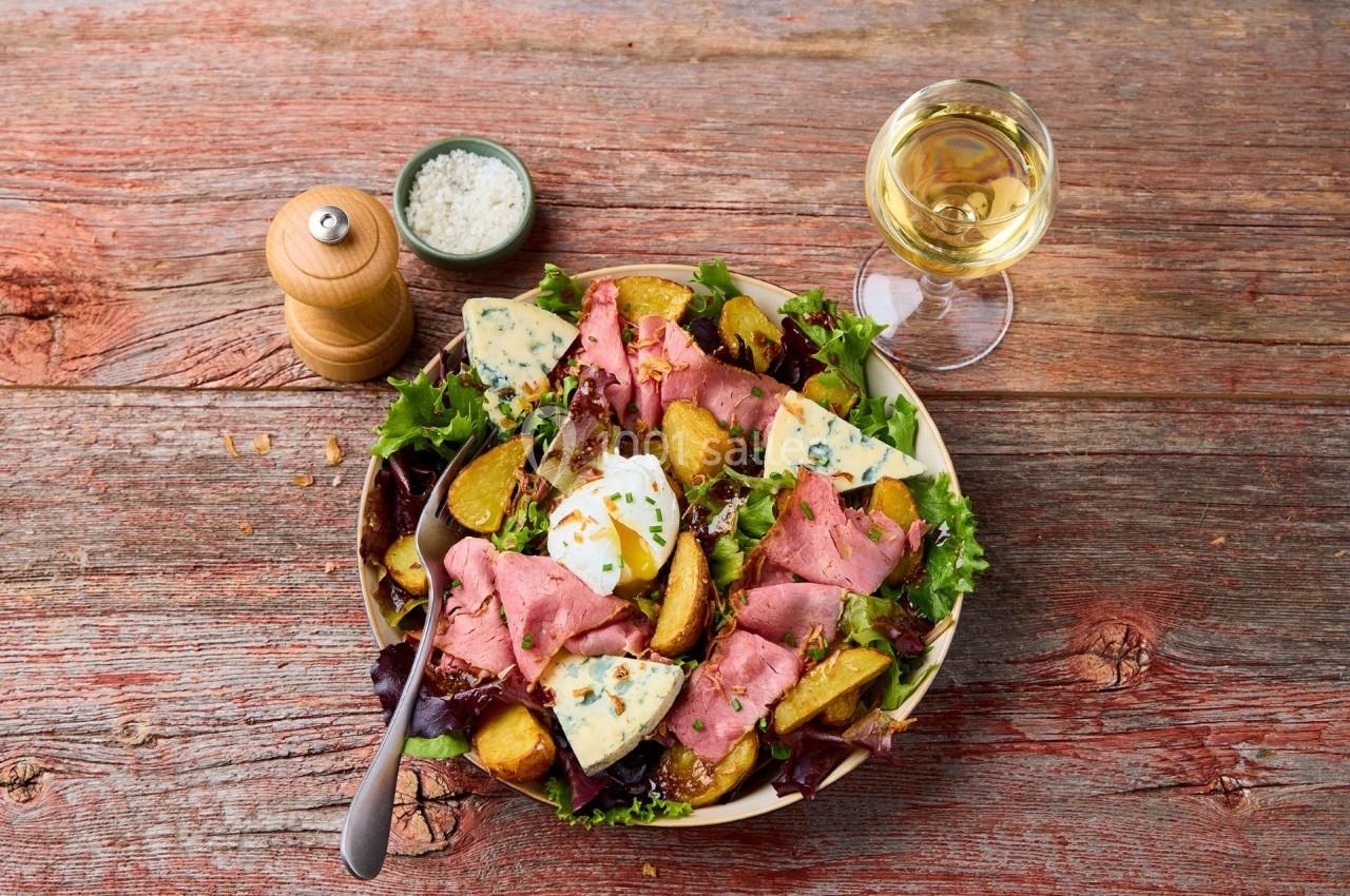 Assiette de salade composée avec rosbif, pommes de terre, fromage bleu, œuf poché et herbes, sur table en bois.