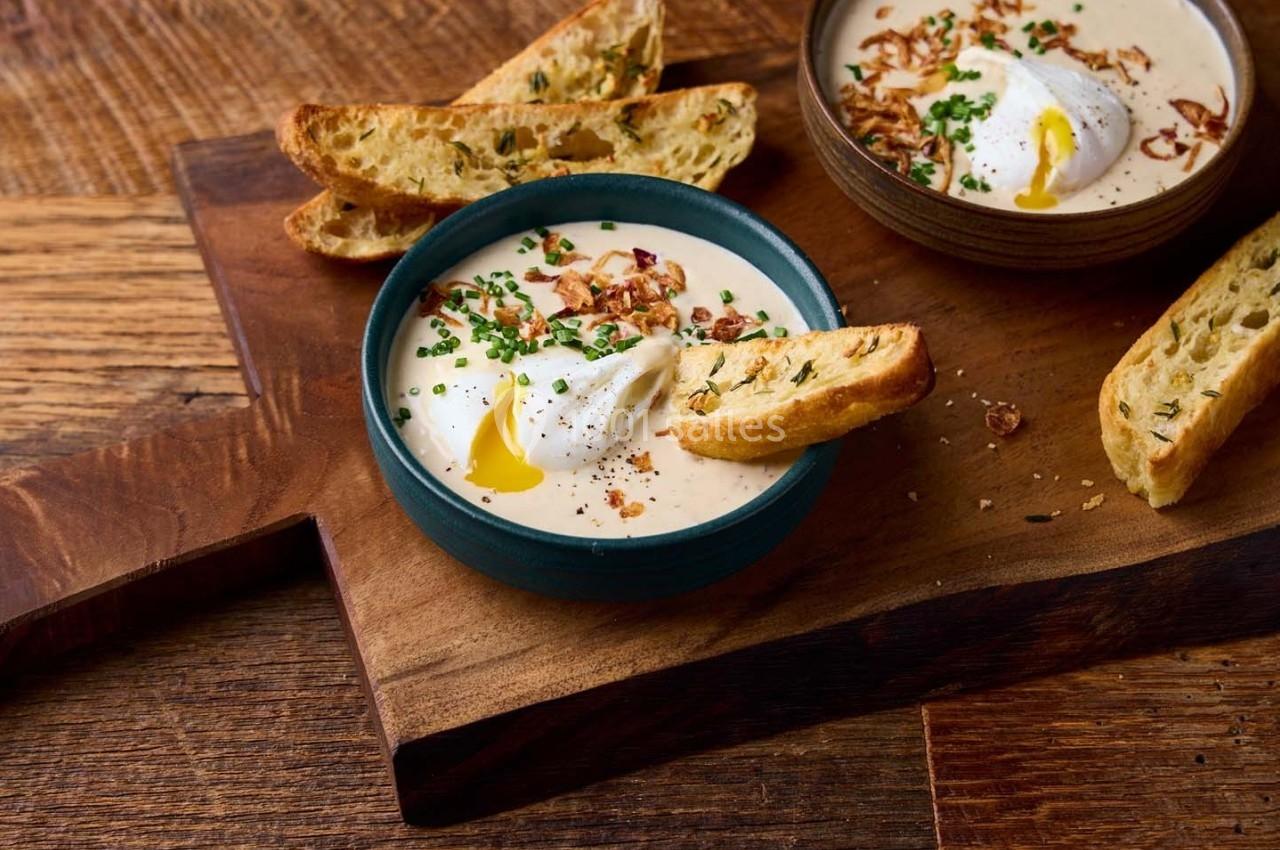 Bol de soupe crémeuse garnie de ciboulette, œufs pochés et croûtons, posé sur une planche en bois.