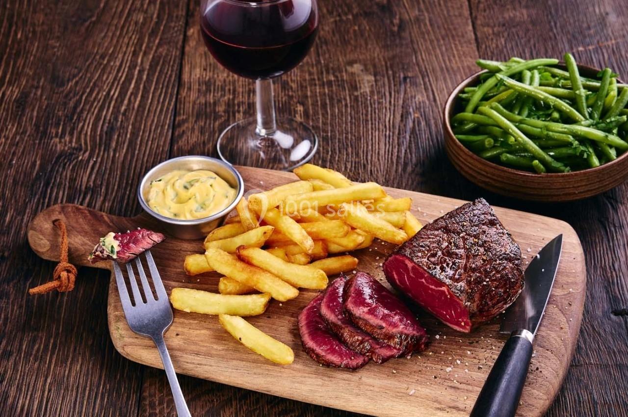 Assiette en bois avec un steak saignant tranché, des frites, une sauce, un verre de vin rouge et des haricots verts.