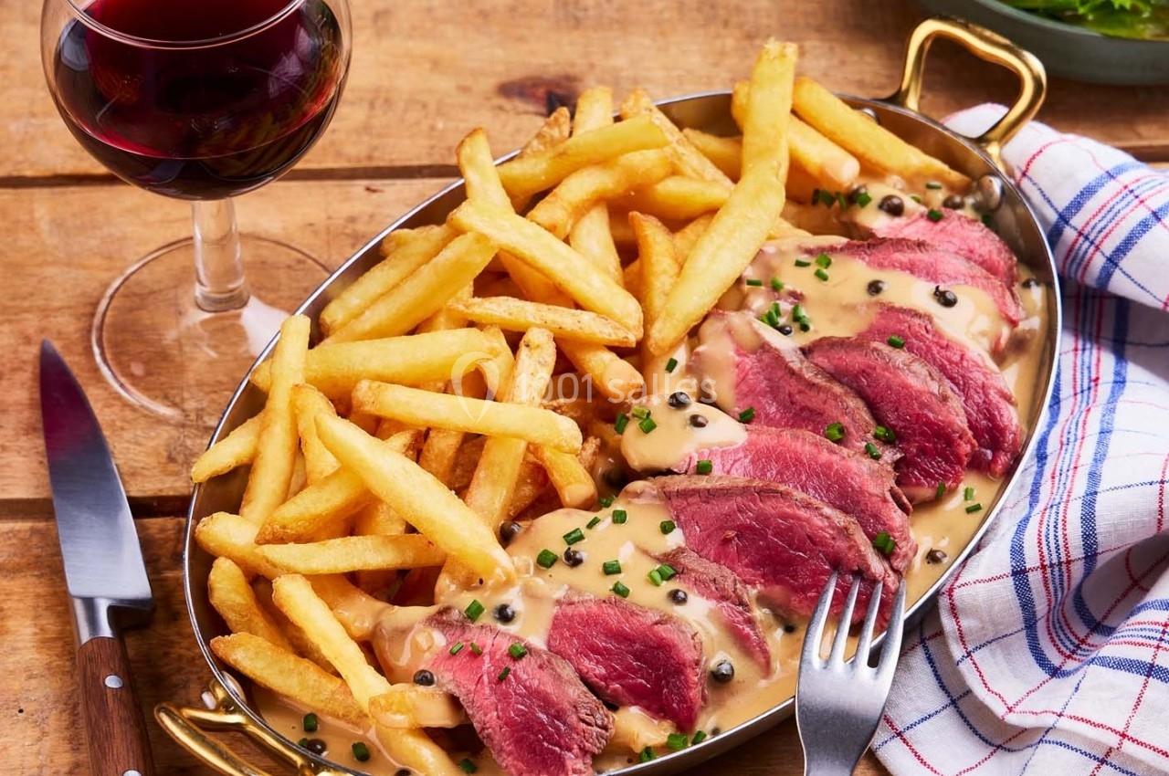 Plat de viande rouge tranchée avec sauce au poivre, accompagné de frites, servi sur un plateau avec un verre de vin rouge.