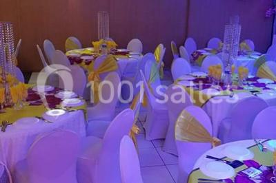 Salle de réception décorée avec des tables rondes dressées de nappes jaunes et rouges, chaises blanches et lustres suspendus.