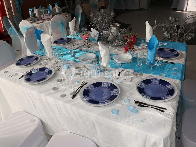 Table dressée avec nappe blanche, chemins de table bleus, assiettes décorées, couverts, serviettes pliées et décorations.