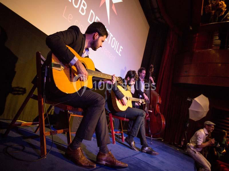 Musiciens jouant de la guitare et de la contrebasse sur une scène éclairée, devant un public dans une salle sombre.