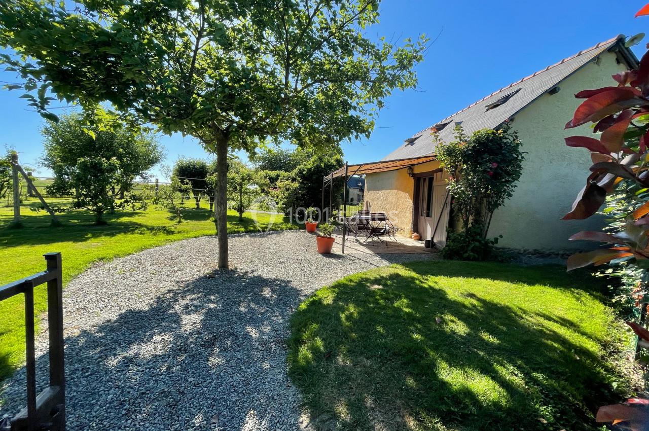 Jardin avec pelouse, arbre, terrasse ombragée et maison à toit en tuiles sous un ciel bleu.