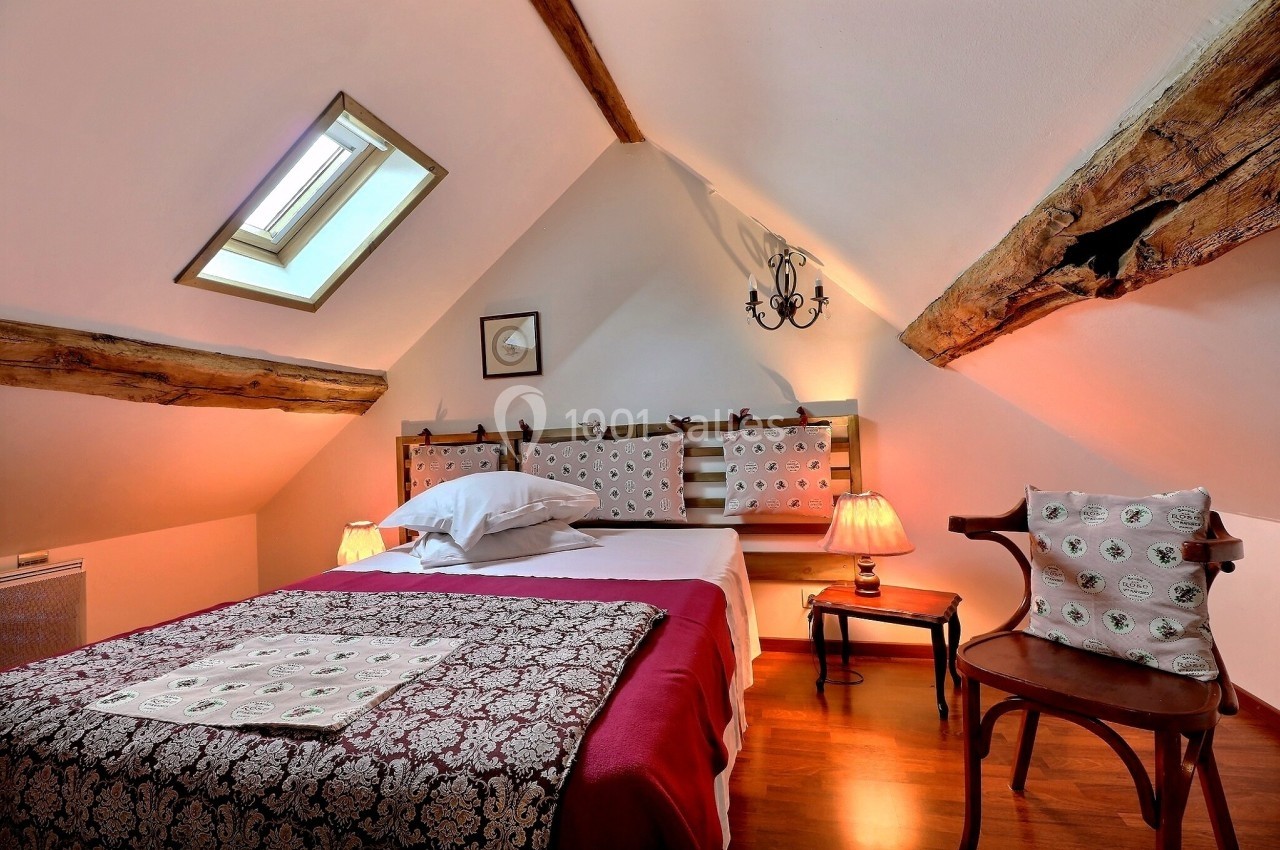 Chambre mansardée avec lit double, poutres apparentes, éclairage chaleureux et fenêtre de toit.