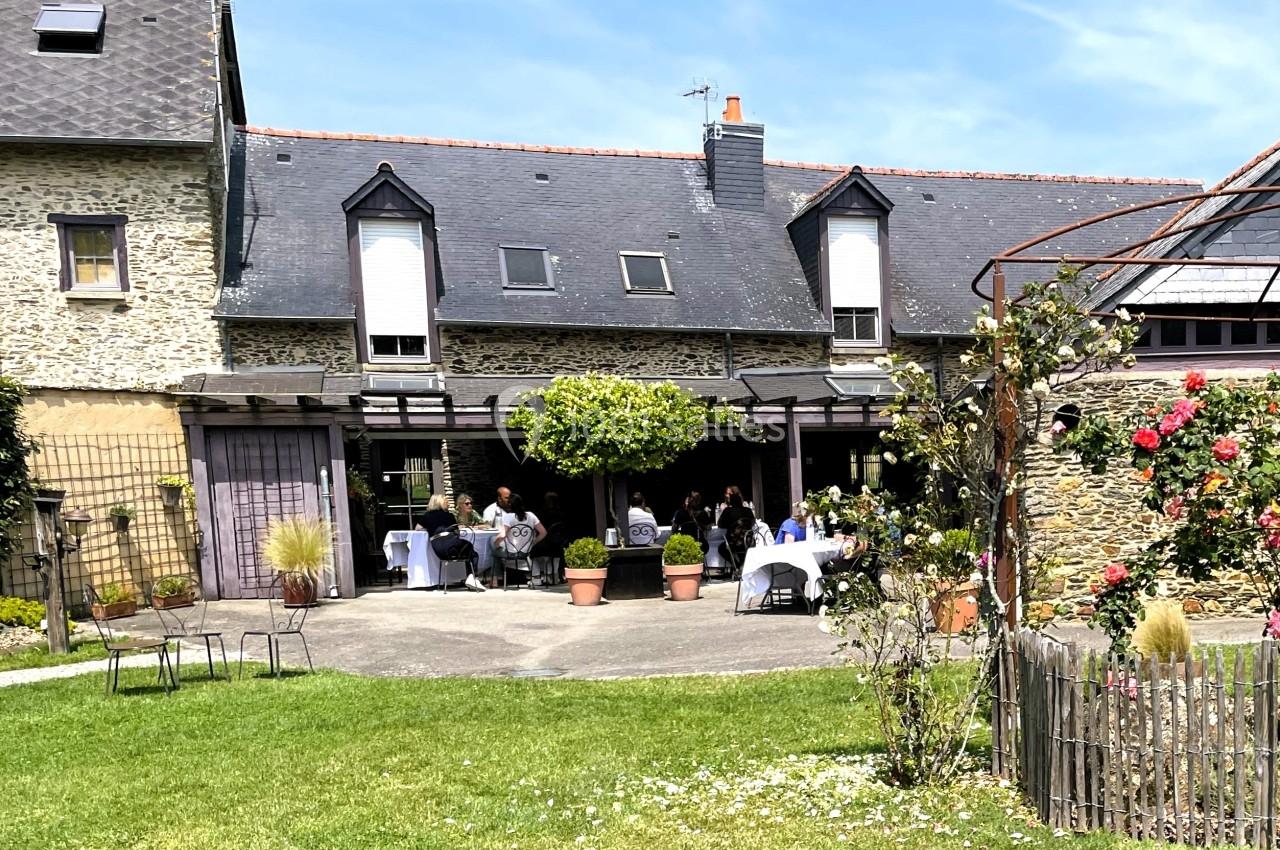 Terrasse d'une maison en pierre avec des tables dressées, entourée de verdure et de fleurs sous un ciel bleu.