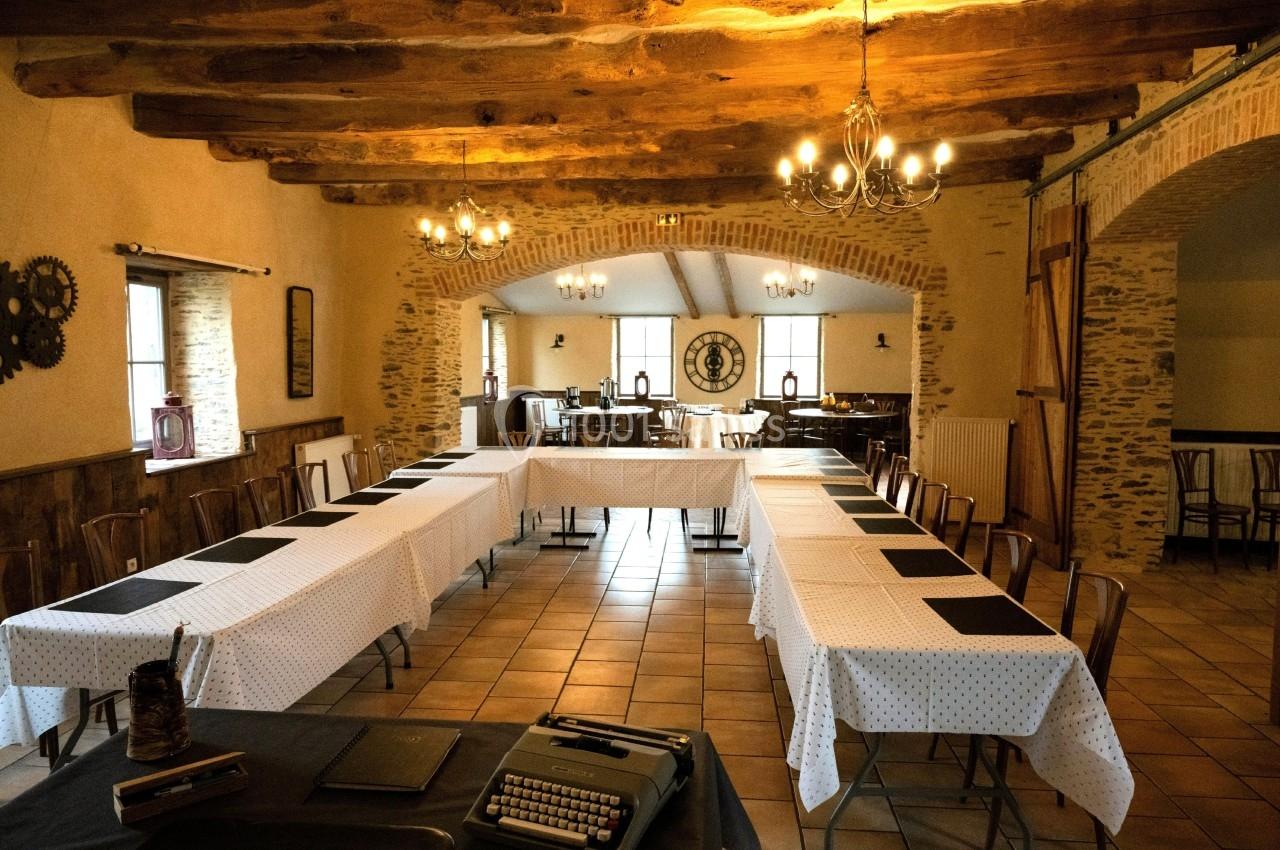 Salle de réunion rustique avec tables disposées en U, nappes blanches et poutres apparentes au plafond.