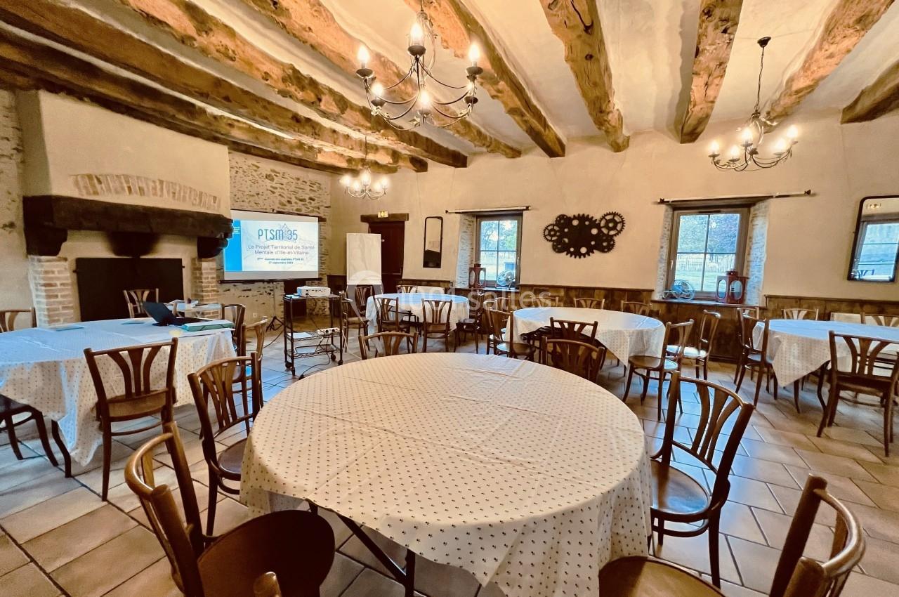 Salle de réunion rustique avec tables rondes couvertes de nappes blanches, poutres apparentes et cheminée en pierre.