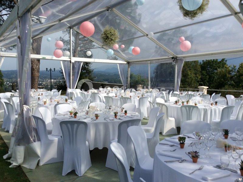 Salle de réception en plein air sous une tente transparente, avec tables rondes décorées et chaises blanches.