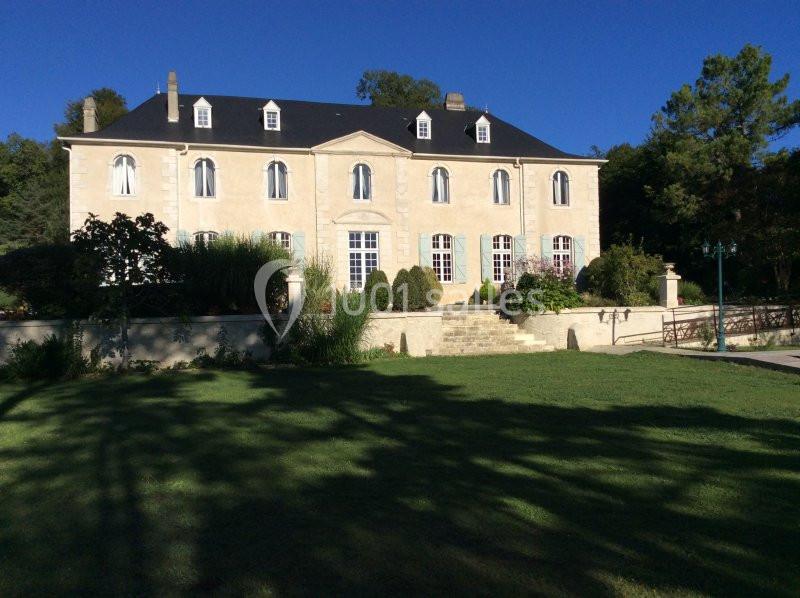 Façade d'une grande maison en pierre avec toit en ardoise, entourée de pelouse et de végétation sous un ciel bleu.
