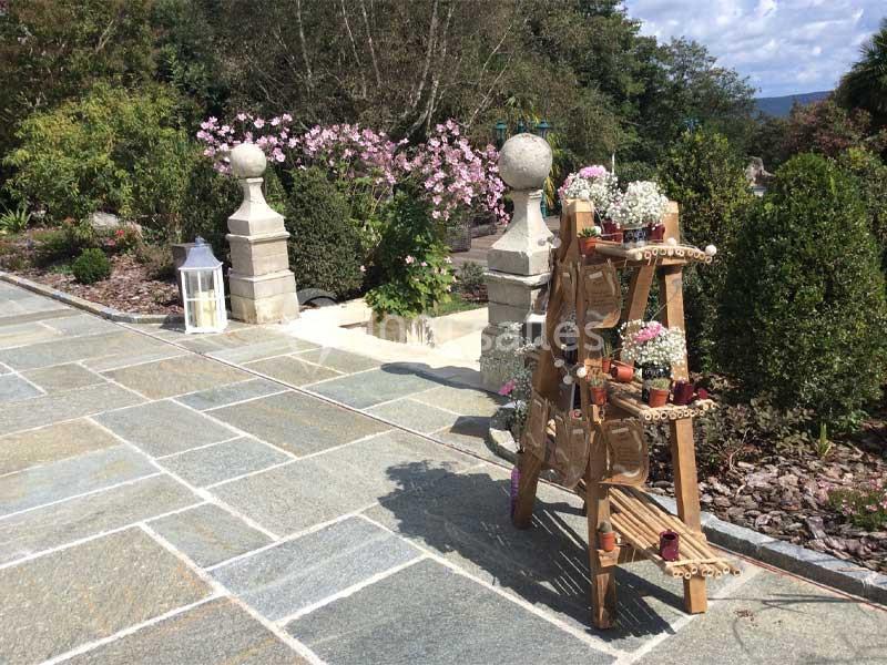 Un présentoir en bois avec des plantes fleuries sur une terrasse en pierre, entourée de verdure et de fleurs.