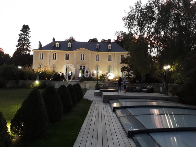 Manoir éclairé au crépuscule, entouré de jardins, avec une terrasse en bois et une piscine couverte au premier plan.