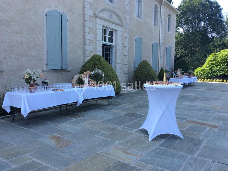 Tables dressées pour un événement en extérieur devant une grande bâtisse avec volets bleus et arbustes taillés.