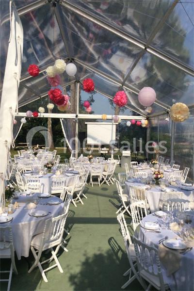 Salle de réception en plein air sous une tente transparente, décorée de lanternes et pompons colorés, tables dressées.