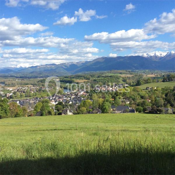 Vue d'un village entouré de collines verdoyantes avec des montagnes en arrière-plan sous un ciel partiellement nuageux.