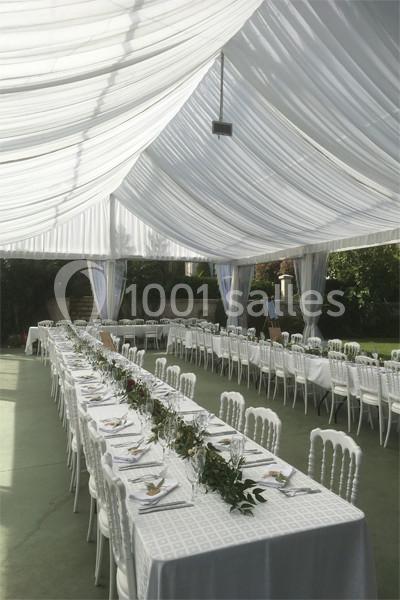 Tables longues dressées avec nappes blanches et décorations végétales sous une tente blanche translucide.