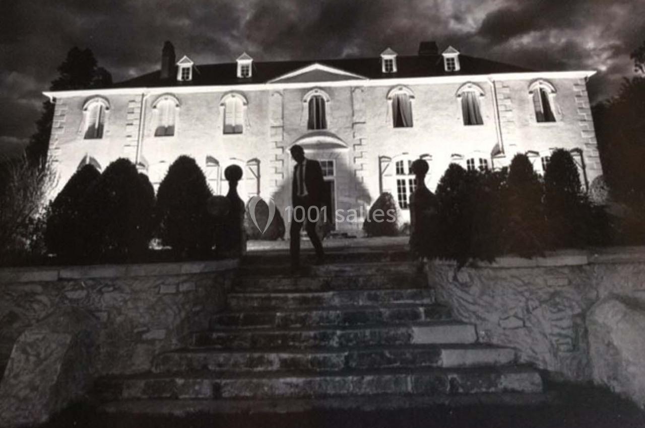 Un homme se tient devant un grand bâtiment éclairé de nuit, entouré de buissons et d'escaliers en pierre.