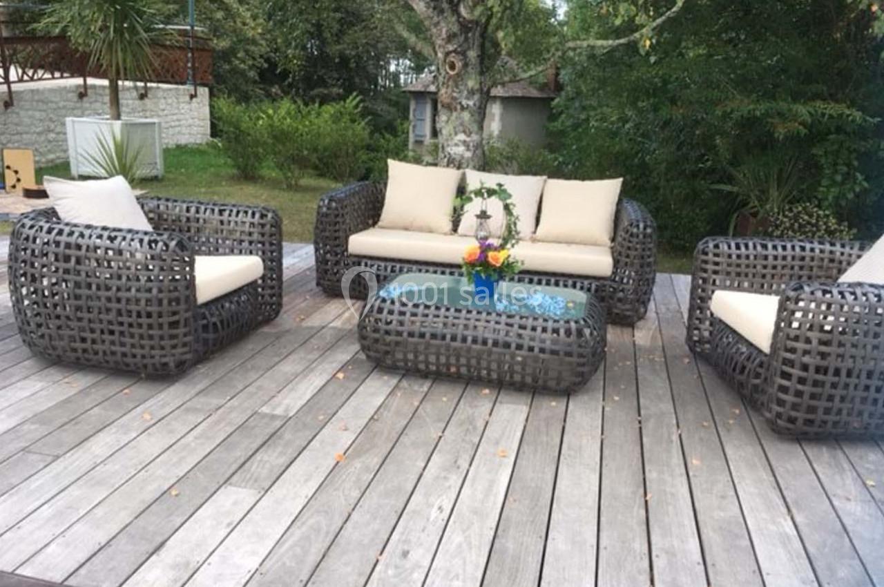 Mobilier de jardin en résine tressée avec coussins beiges, disposé sur une terrasse en bois entourée de verdure.