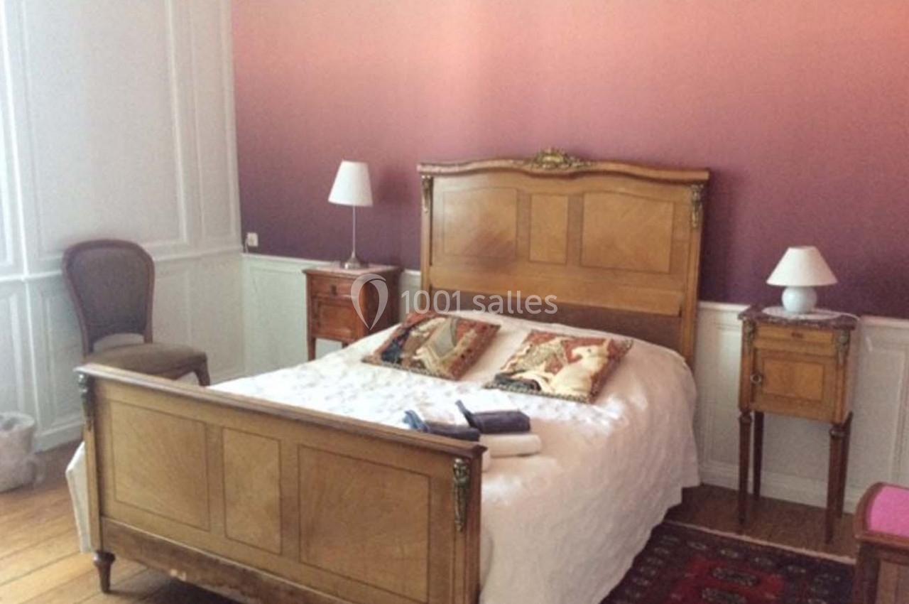 Chambre avec lit en bois ancien, murs dégradés rose-violet, deux tables de chevet et une chaise en bois.