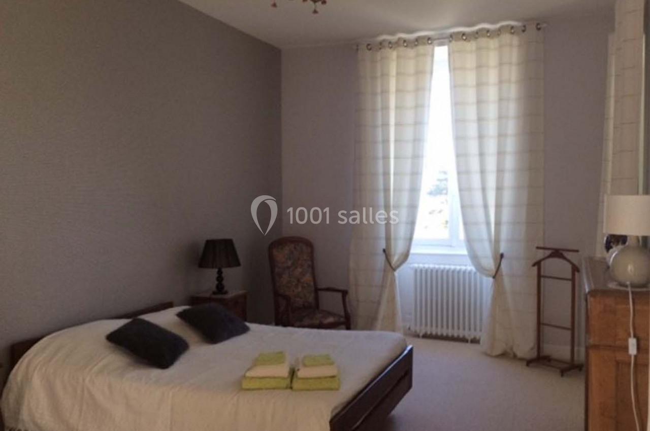 Chambre lumineuse avec lit double, rideaux blancs, fauteuil, lampe de chevet et décoration sobre.