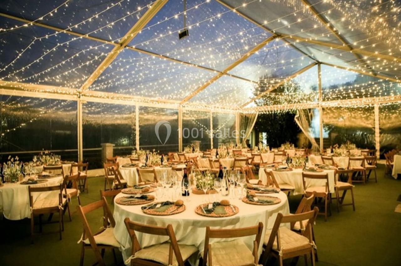 Salle de réception en soirée sous une tente transparente, décorée de guirlandes lumineuses et tables dressées.