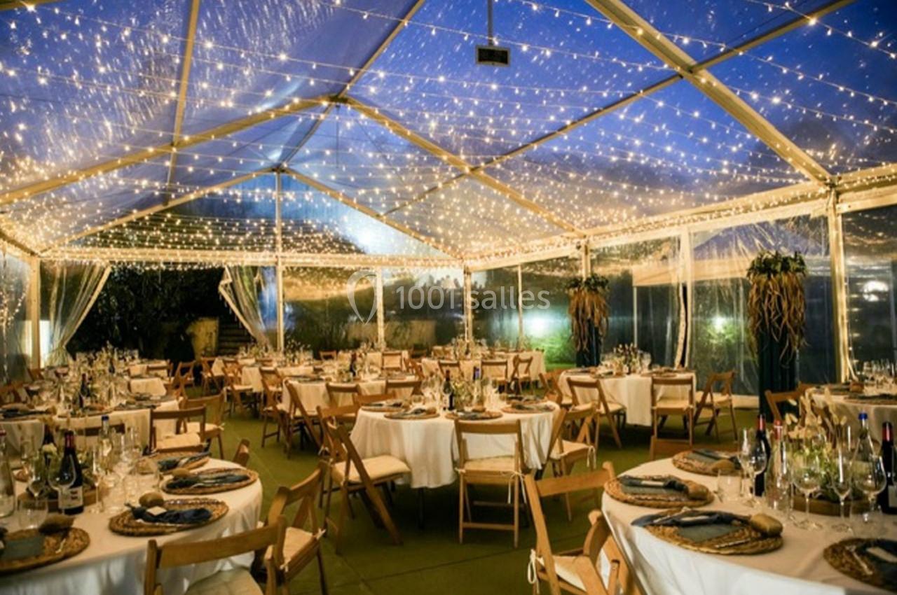 Salle de réception sous une tente transparente illuminée par des guirlandes, avec tables dressées pour un événement.