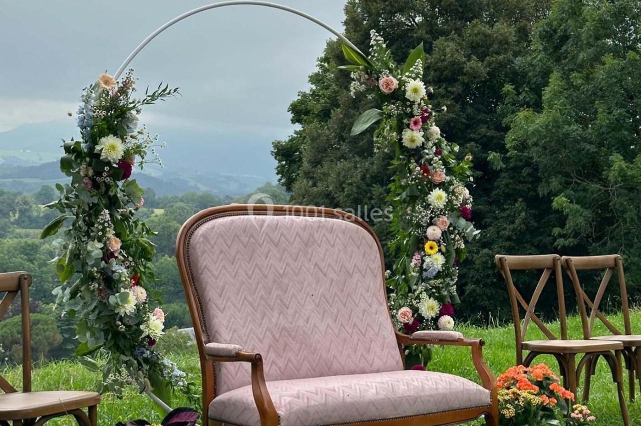 Arche florale décorée avec des fleurs colorées, fauteuil rose et chaises en bois dans un cadre naturel verdoyant.