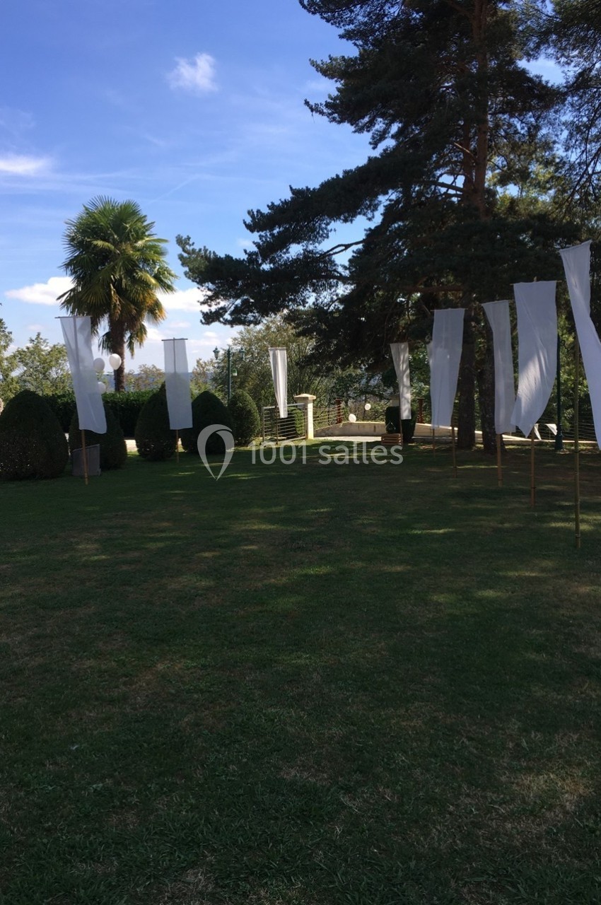 Des drapeaux blancs verticaux flottent dans un jardin herbeux entouré d'arbres sous un ciel bleu.