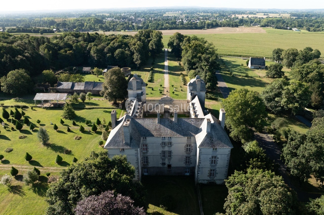 Vue aérienne d'un château entouré de jardins paysagers, de pelouses et d'arbres, avec une campagne en arrière-plan.